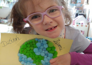 dziewczynka w okularach pokazuje swoja pracę: kontur Ziemi wypełniony miękkimi, małymi zielono - niebieskimi kulkami